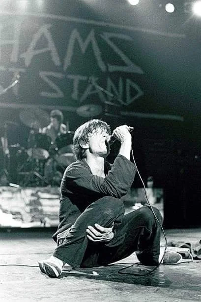 Jimmy Pursey crouched on stage with a microphone, a Sham's Last Stand backdrop behind him