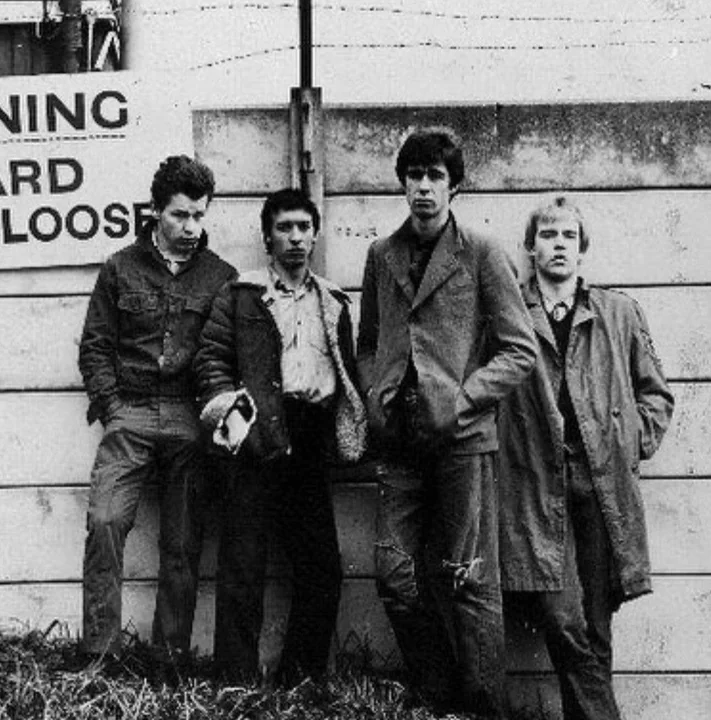 A black and white photograph of a punk group by a street hoarding